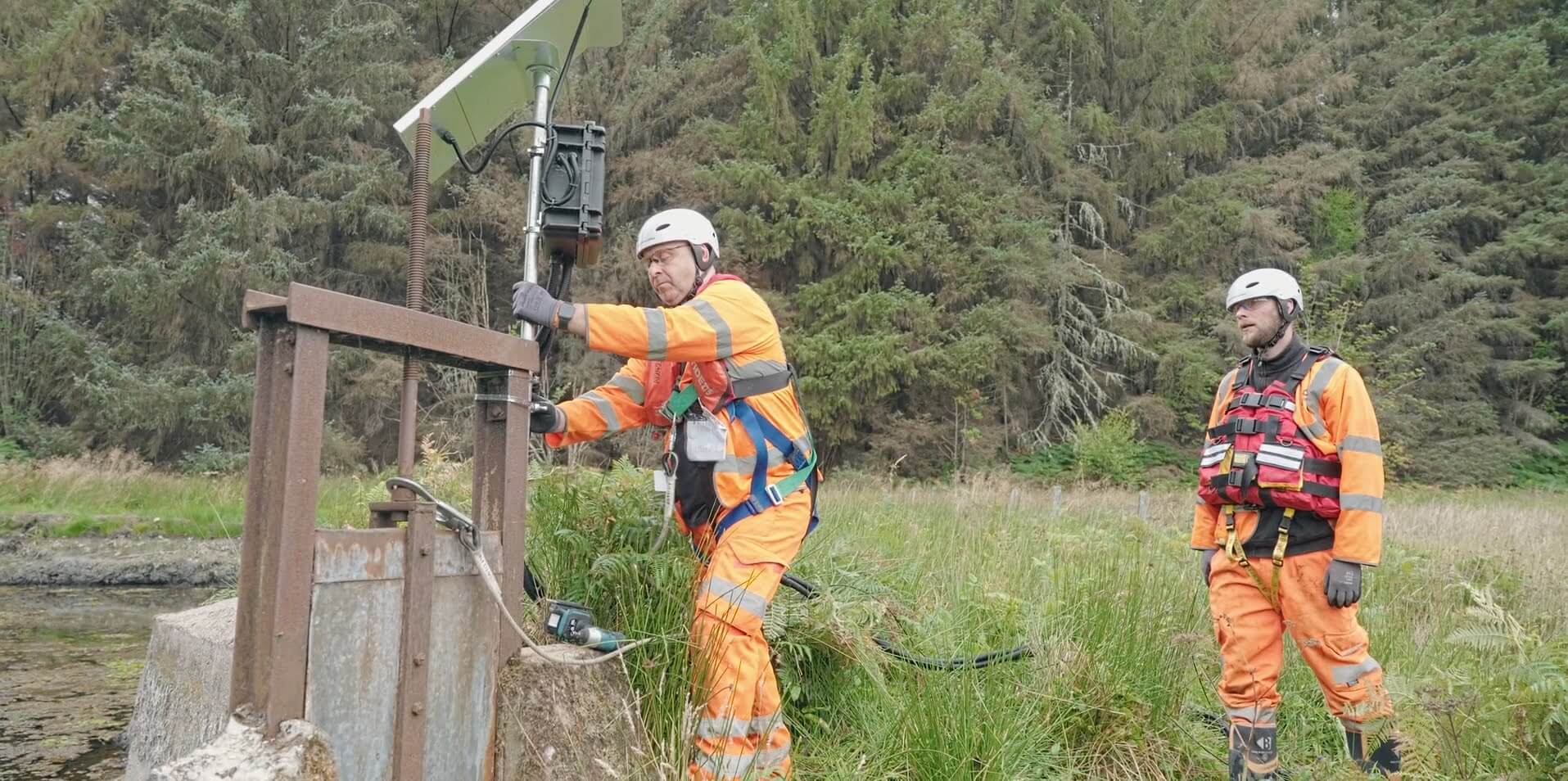 Two workers in high-visibility gear installing a solar-powered water monitoring device beside a rural stream.